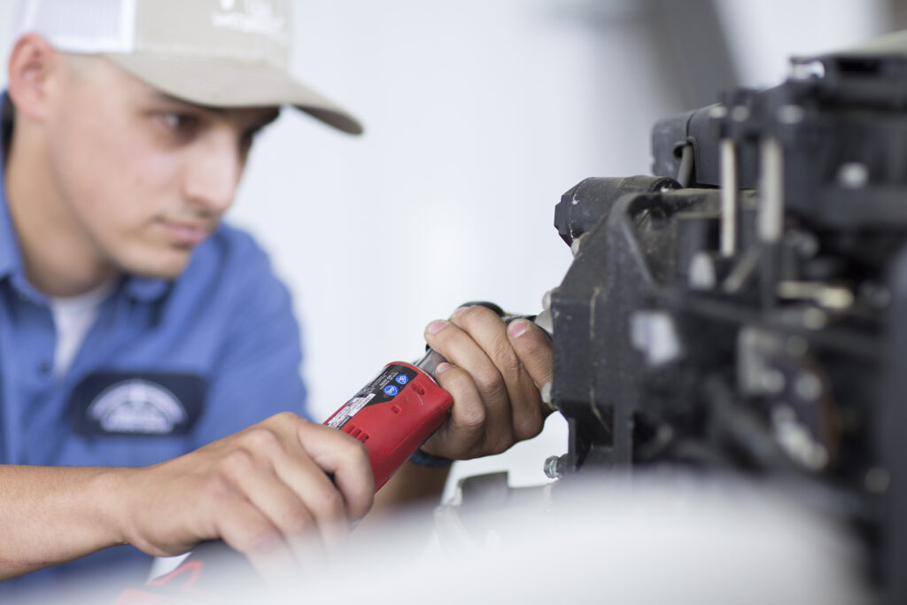 A focused young man in a blue work shirt and beige cap uses a red power tool to work on a piece of marine equipment, highlighting his dedication and technical skills in the marine industry.